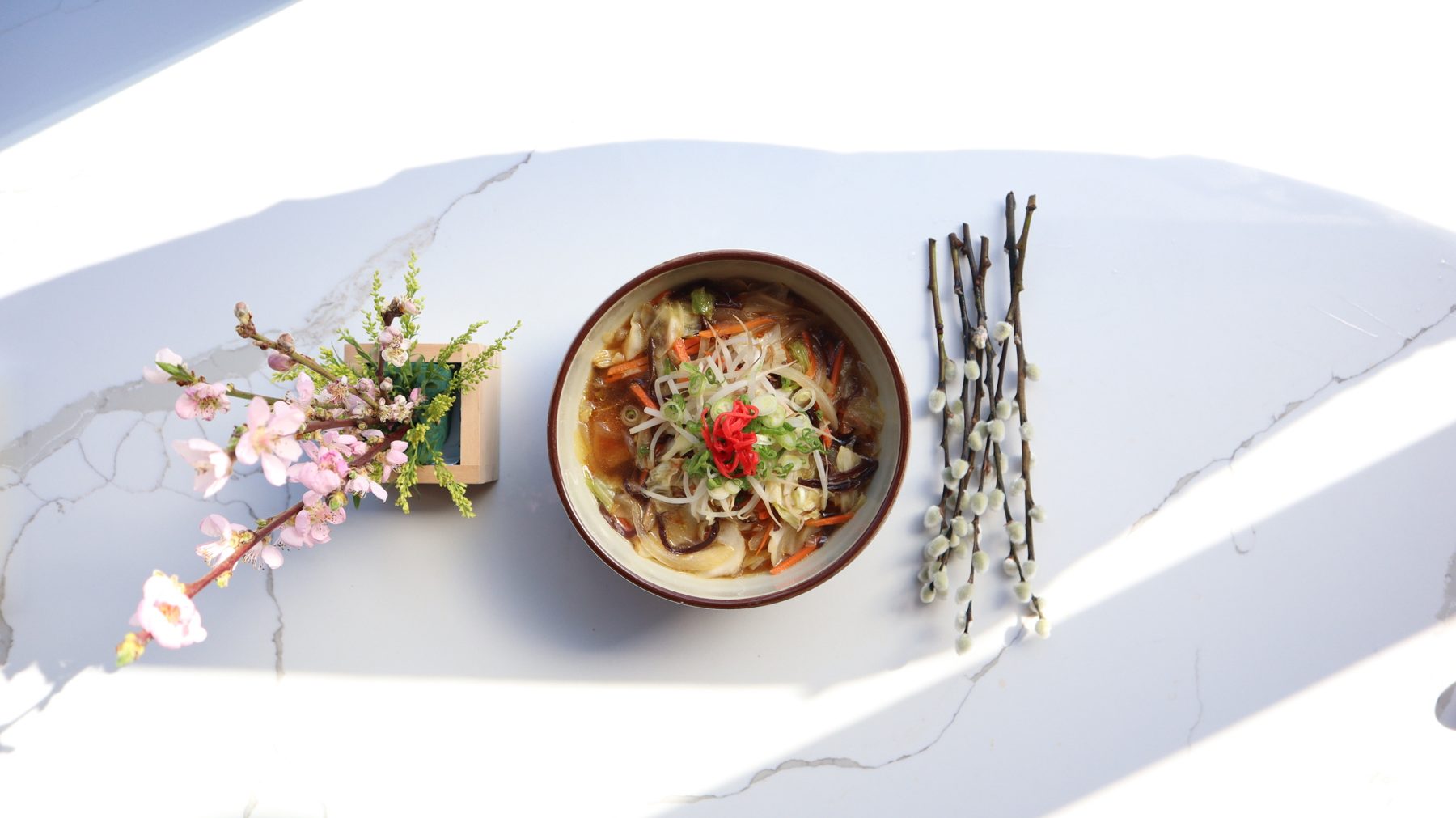 A bowl of noodle soup on a marble table beside peach blossoms and pussy willow